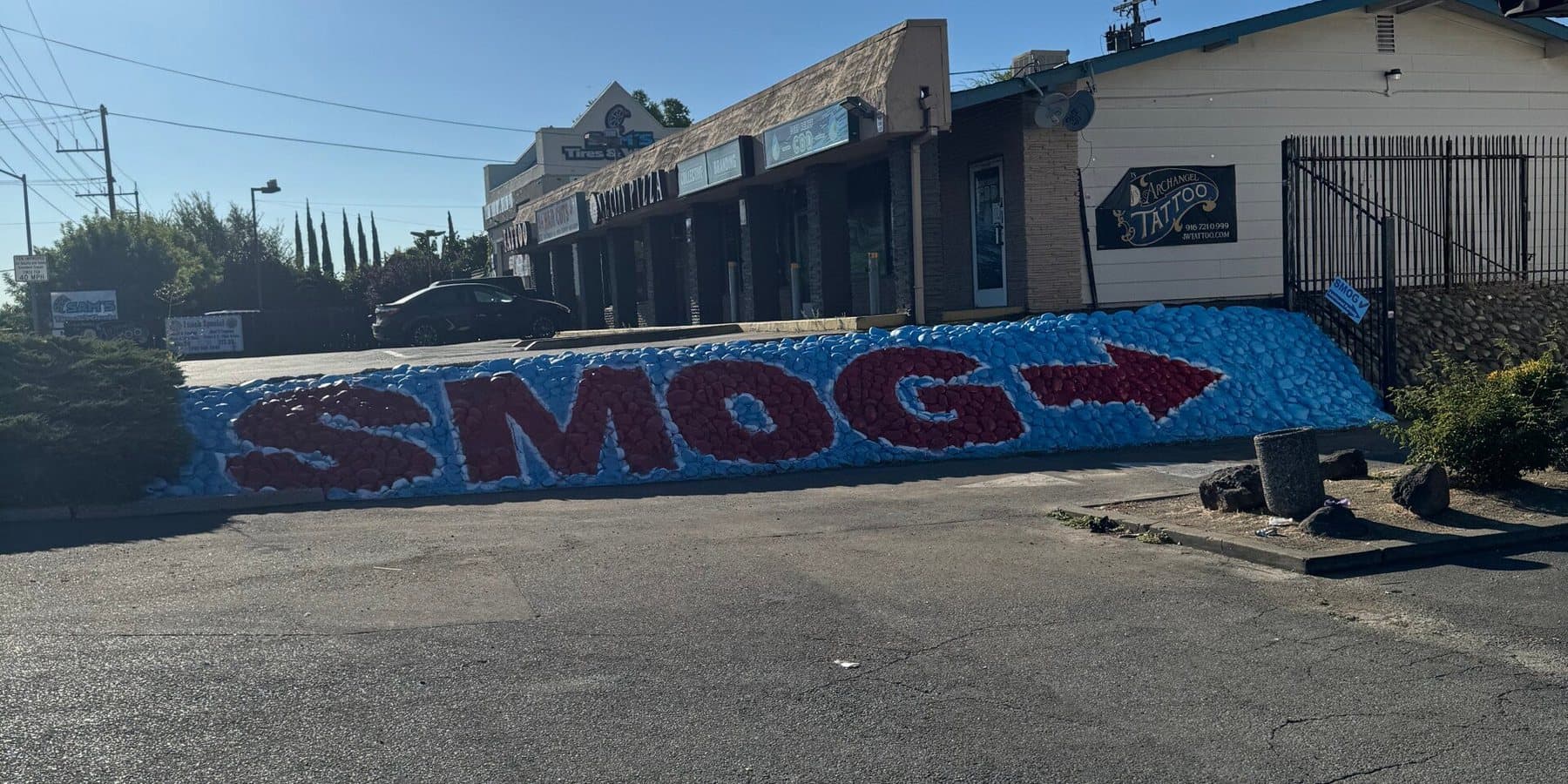Painted rock wall on Greenback Lane spelling SMOG, with a red arrow pointing along the plaza toward the shop