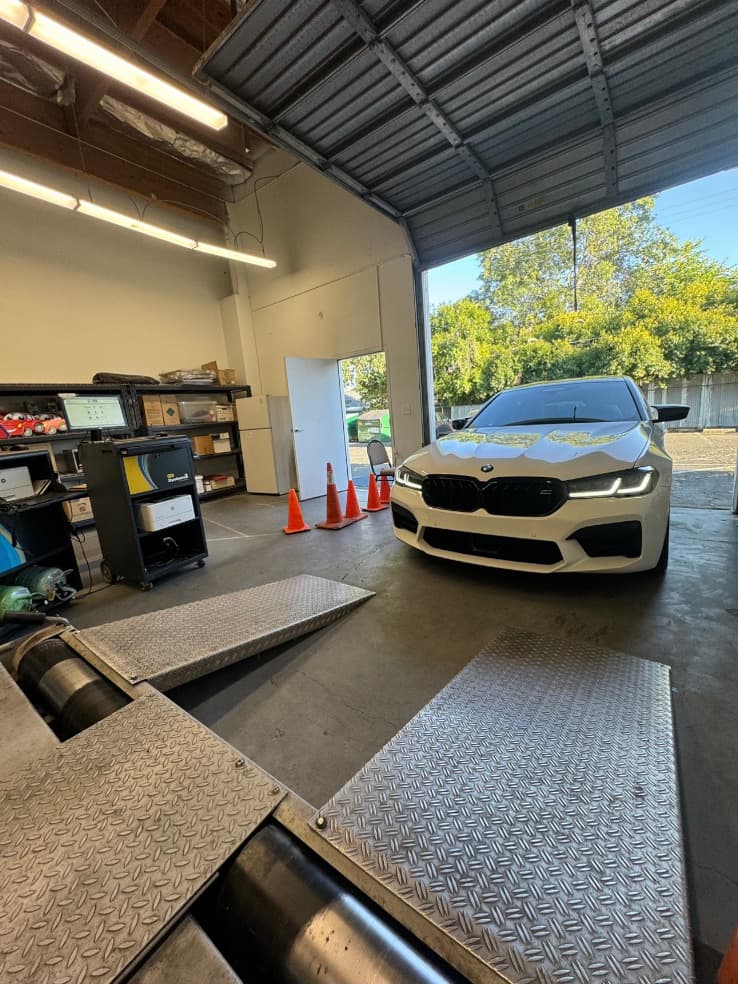 White BMW parked inside the inspection bay by the open roll-up door, facing the dynamometer ramps