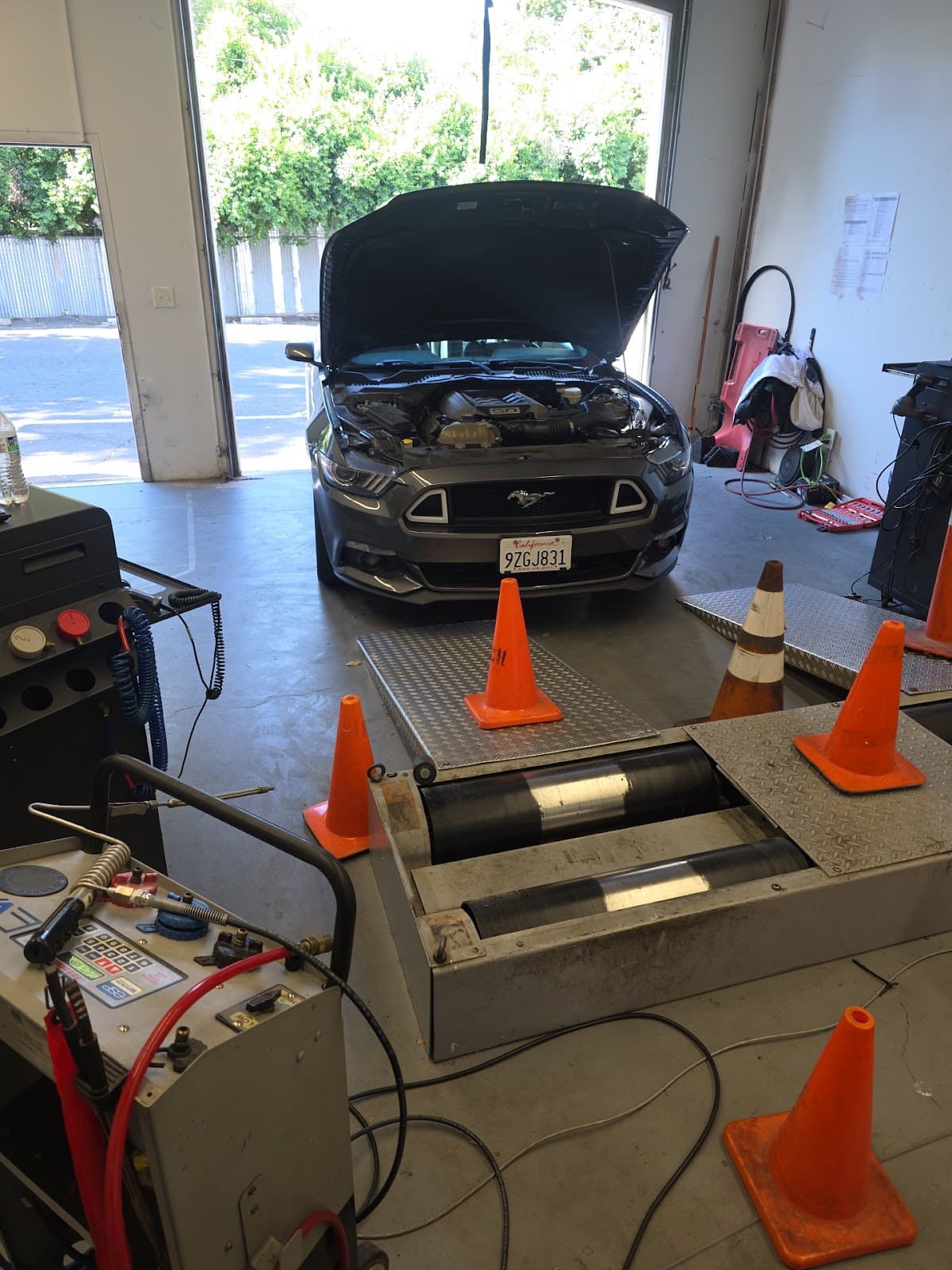 Ford Mustang with its hood raised, parked behind the dynamometer rollers and traffic cones in the inspection bay