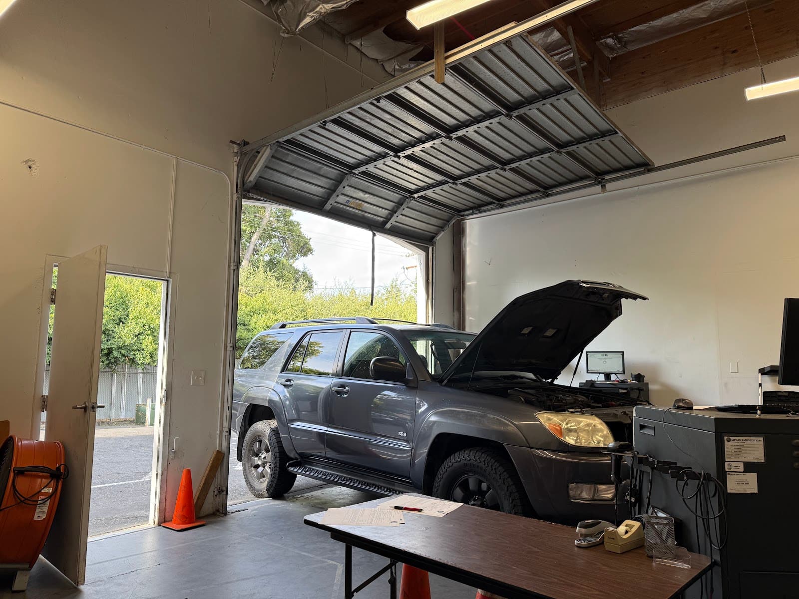 SUV with its hood raised in the shop's inspection bay next to the emissions analyser, with the roll-up door open
