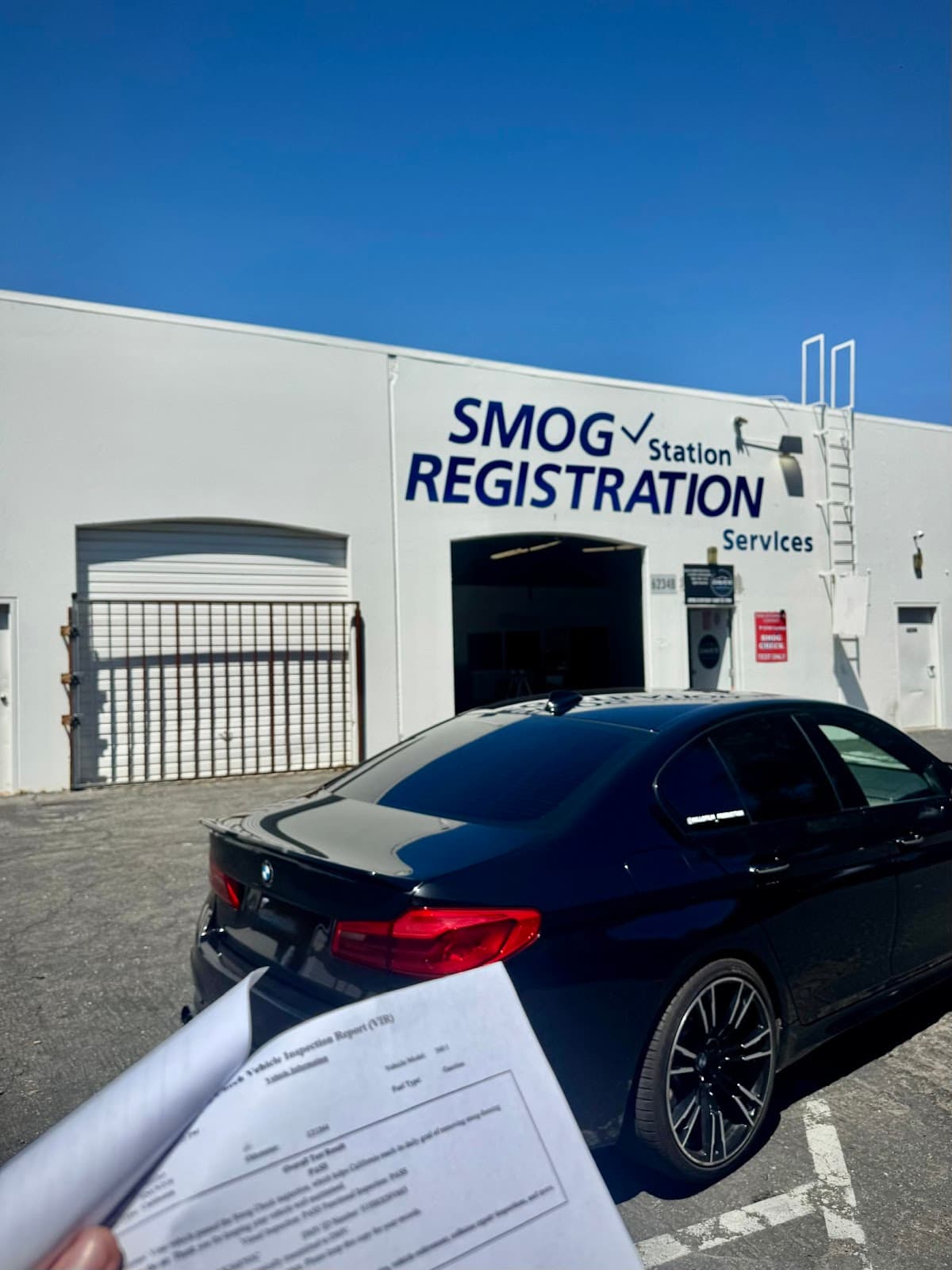 Vehicle inspection report showing a PASS result, held up in front of a BMW parked outside the shop
